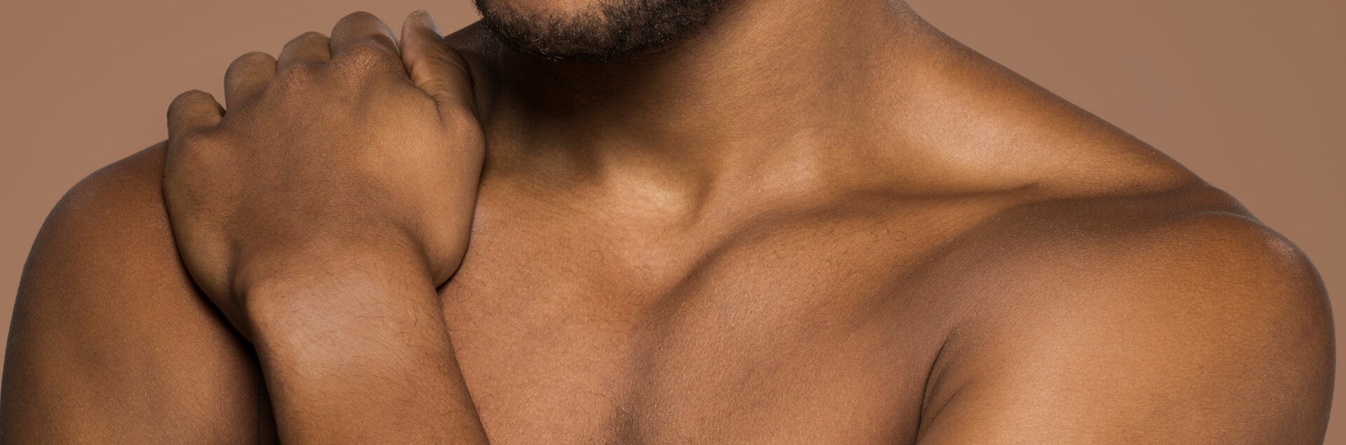 Close-up van de ontblote borst en schouder van een man met een donkere huid. Zijn hand rust op zijn tegenoverliggende schouder en de achtergrond is effen beige.