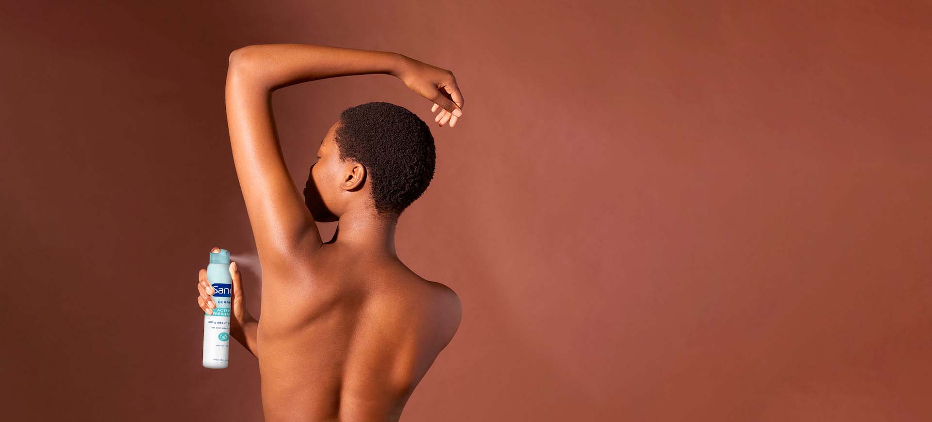 Een zwarte vrouw met kort haar, van achteren gezien, die een spuitbus deodorant onder haar opgestoken arm aanbrengt tegen een bruine achtergrond.
