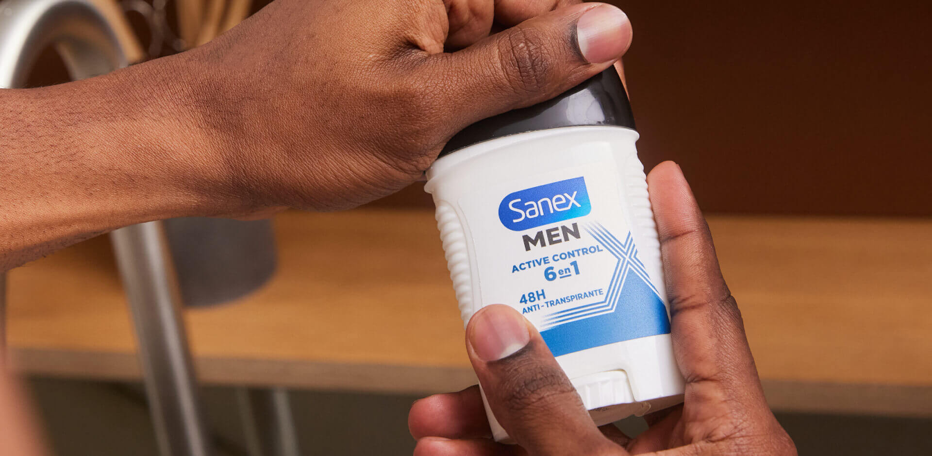 Close-up van de handen van een persoon met een donkere huid, die een witte deodorantstick van Sanex MEN Active Control vasthoudt.