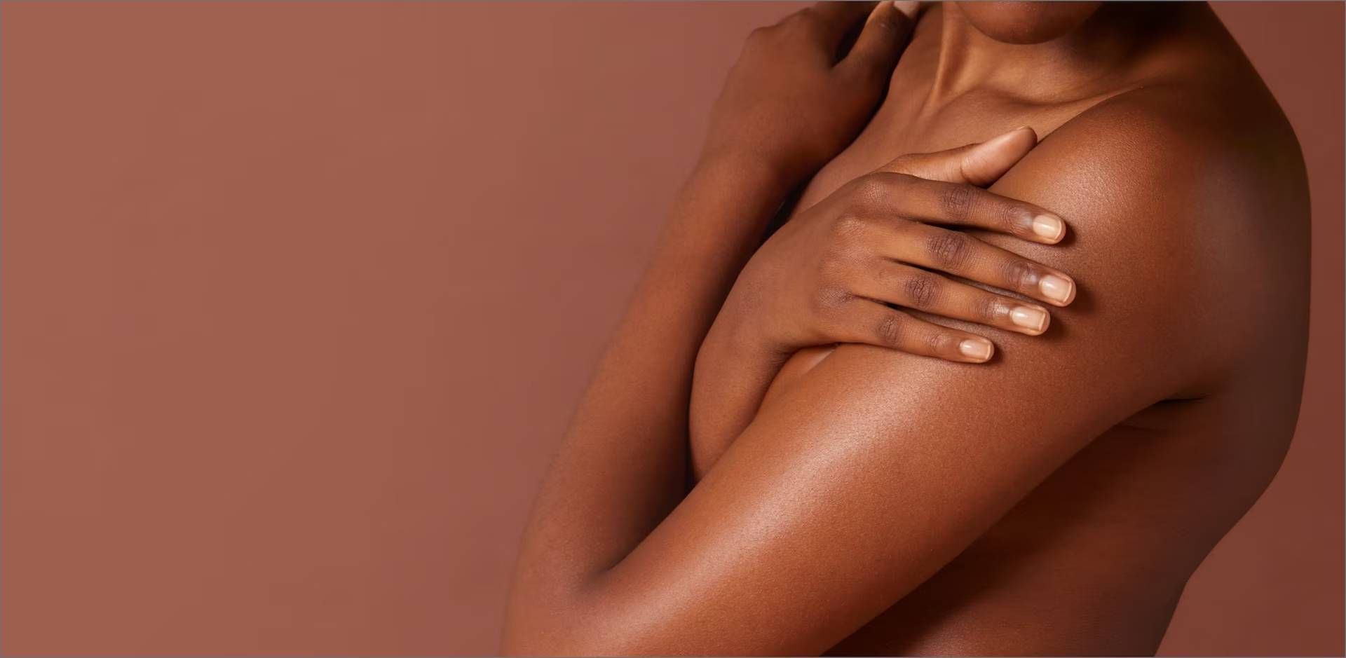 Close-up van een persoon met een donkere huid die zichzelf omarmt, waarbij de hand zachtjes op de schouder rust. De achtergrond is effen bruin.