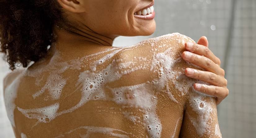 Een lachende vrouw, van achteren gezien, die haar schouder en rug inzeept met een rijk schuim terwijl ze onder de douche staat.