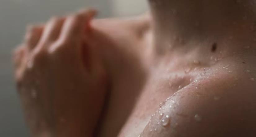 Intieme close-up van een natte schouder die bedekt is met waterdruppels. Op de achtergrond rust een onscherpe hand op de huid.