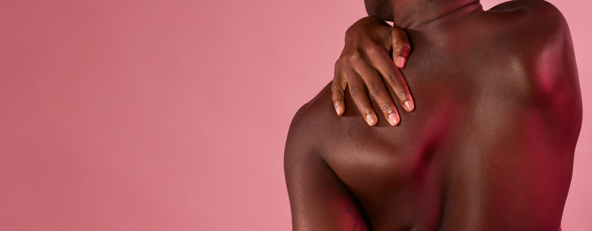 Close-up van de rug van een persoon met een donkere huid, wiens hand op de tegenoverliggende schouder rust. De achtergrond is roze.
