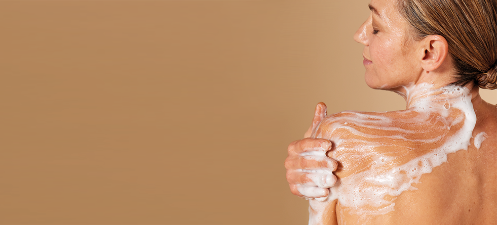 Een vrouw met een lichte huidskleur, gezien vanaf haar rug, die haar schouder inzeept met een rijk schuim tegen een beige achtergrond.