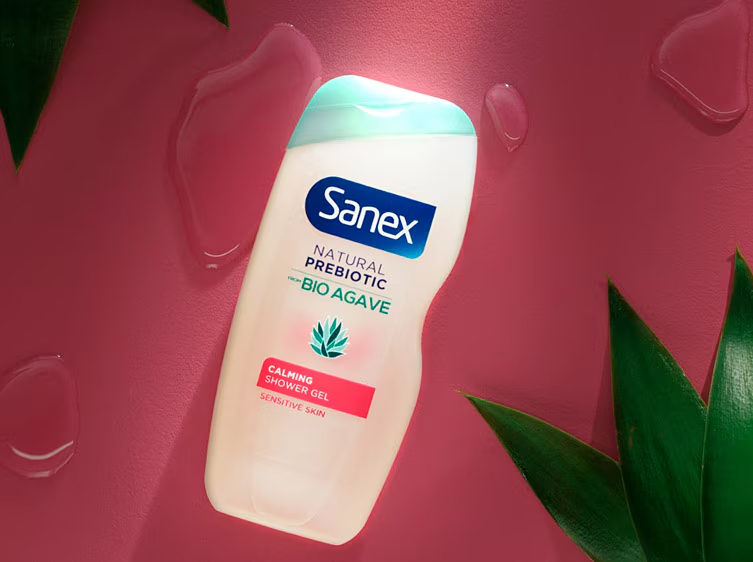 Sanex Natural Prebiotic Bio Agave Calmante Douchegel met roze label op een roze achtergrond met groene bladeren.